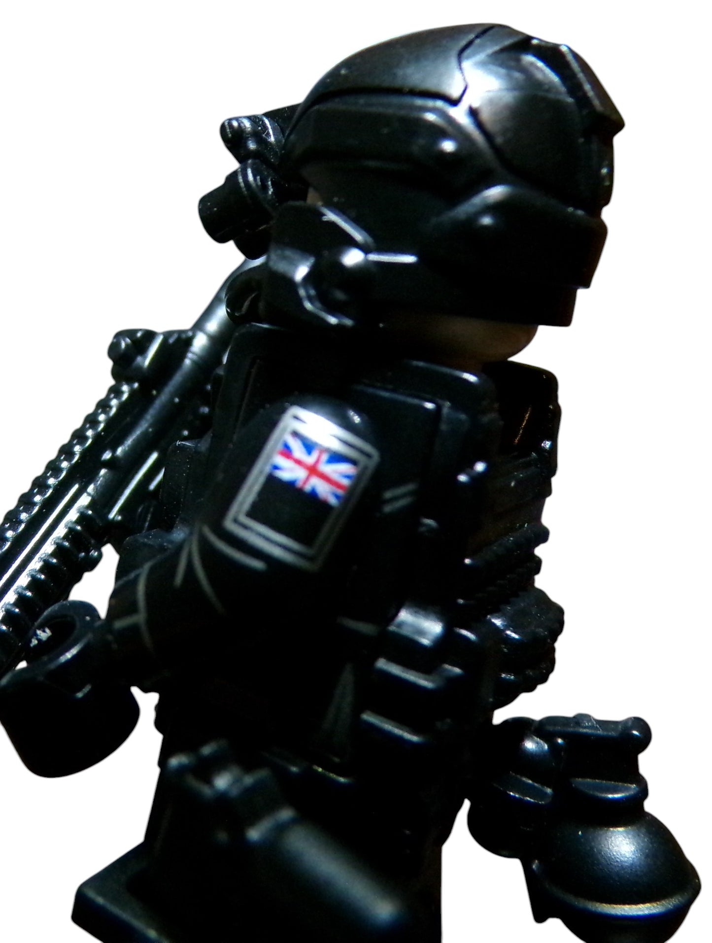 A close-up of a LEGO Special Air Service minifigure in black tactical gear, featuring a helmet, goggles, and a Union Jack on its arm. The figurine has a weapon slung across its back against a plain white background.