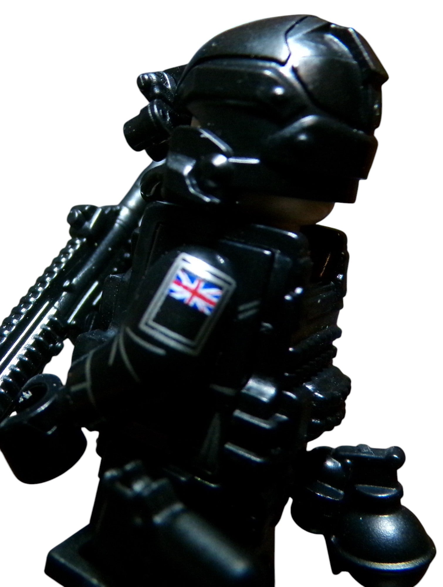 A close-up of a LEGO Special Air Service minifigure in black tactical gear, featuring a helmet, goggles, and a Union Jack on its arm. The figurine has a weapon slung across its back against a plain white background.