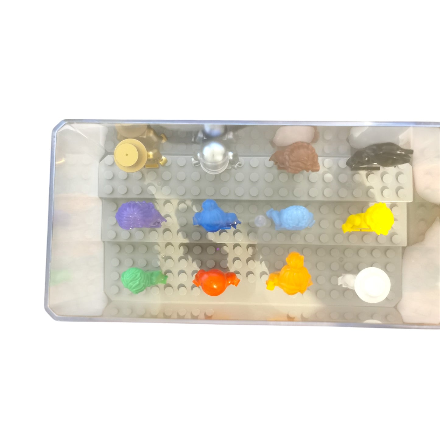 A clear plastic display case presents 12 Vibrant Colour Minifigures on a gray, perforated base. These striking figures resemble animals and objects, showcasing hues of blue, yellow, orange, purple, and green from above.