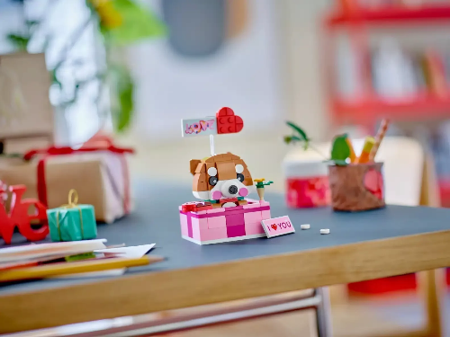 A small desk displays a LEGO® Love Gift Box (40679) featuring a Shiba Inu dog with a heart-shaped hat on a pink and white Valentines Day box reading I love you. Wrapped gifts, pencils, and plants are nearby against blurred background shelves.