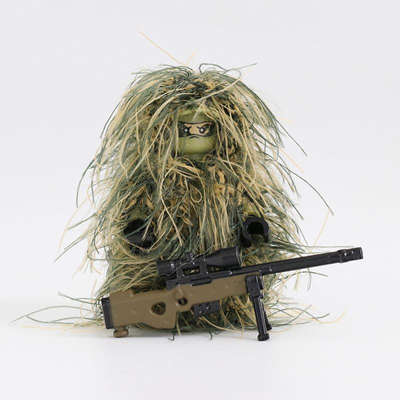 The Custom LEGO Minifigure Sniper, camouflaged in a green and brown ghillie suit, holds a toy rifle. Its face is partially hidden behind dark glasses on a plain white backdrop.