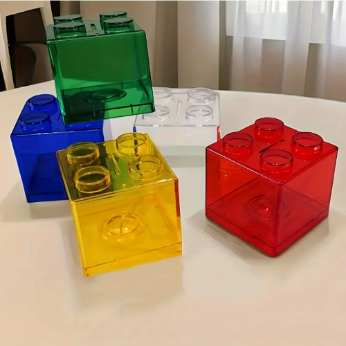On the table are four large, transparent Building Block Piggy Banks in green, blue, yellow, and red. Theres also a clear block—ideal for inspiring saving habits with its playful design resembling oversized building bricks.