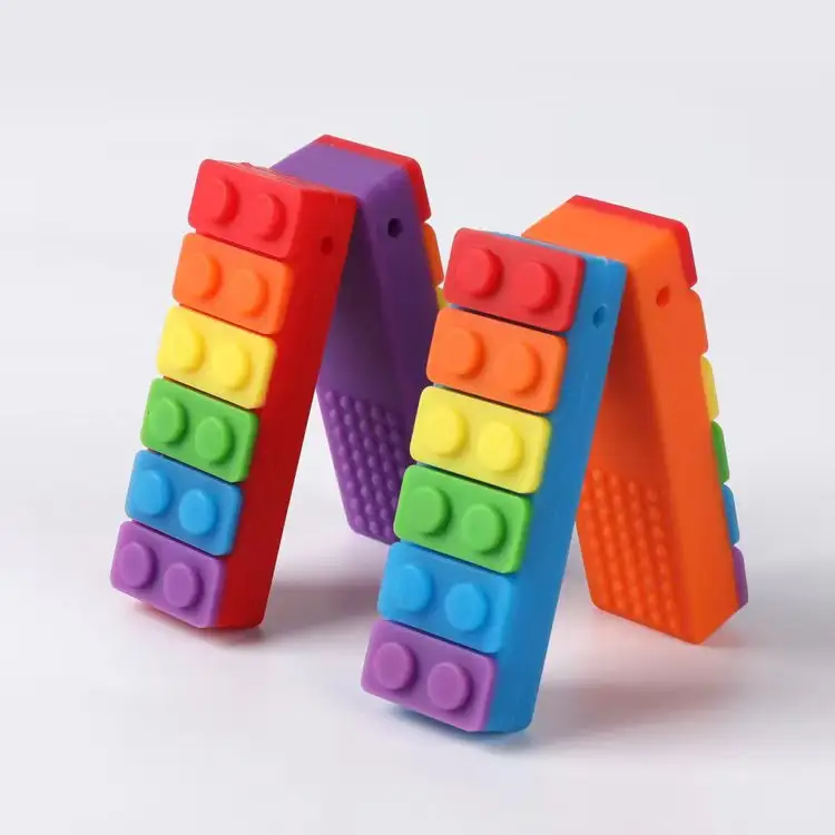 Two vibrant toy bridges of interlocking blocks mimic the Sensory Chew Necklace Rainbow Chew Toys, showcasing red, orange, yellow, green, blue, and purple in a graduated pattern. These stand on a white surface with a minimalist backdrop.
