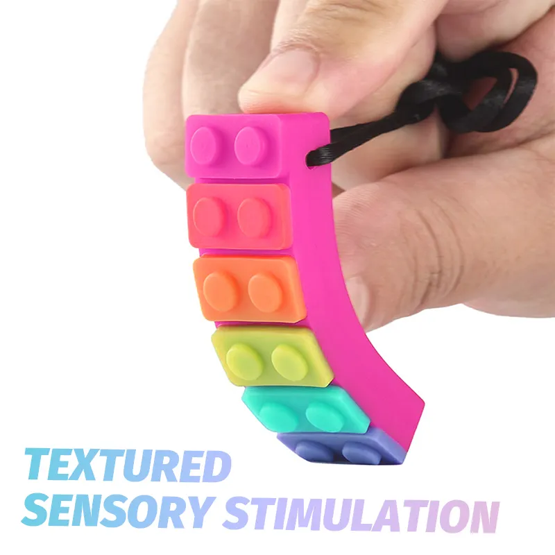A hand holds a vibrant Sensory Chew Necklace Rainbow Chew Toy, featuring stacked, interlocking blocks in pink, red, orange, yellow, green, and blue. Text below reads TEXTURED SENSORY STIMULATION, highlighting its function as an oral sensory aid for children with sensory needs.