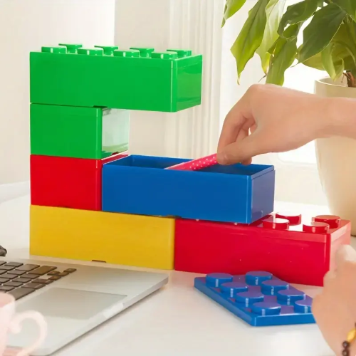 A person is assembling a Pack of 4 2x4 Storage Blocks in red, yellow, blue, and green on a table near a laptop, with the vibrant pieces contrasting against a lush potted plant in the background.