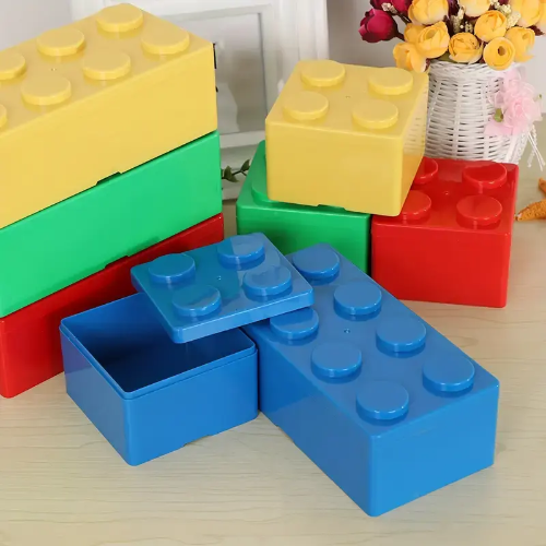 A Pack of 4 2x2 Building Block Storage Containers in red, blue, yellow, and green are stacked on a table. One blue container is open. A bouquet of flowers in a white wicker basket sits gracefully in the background.