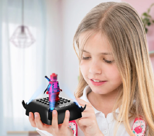 A young girl with long blonde hair smiles, admiring a small holographic block figure glowing blue and red on its 10 Color Changing Lighting Base for Minifigures. The softly lit interior with a hanging light fixture and blurred decor enhances the color-changing ambiance.
