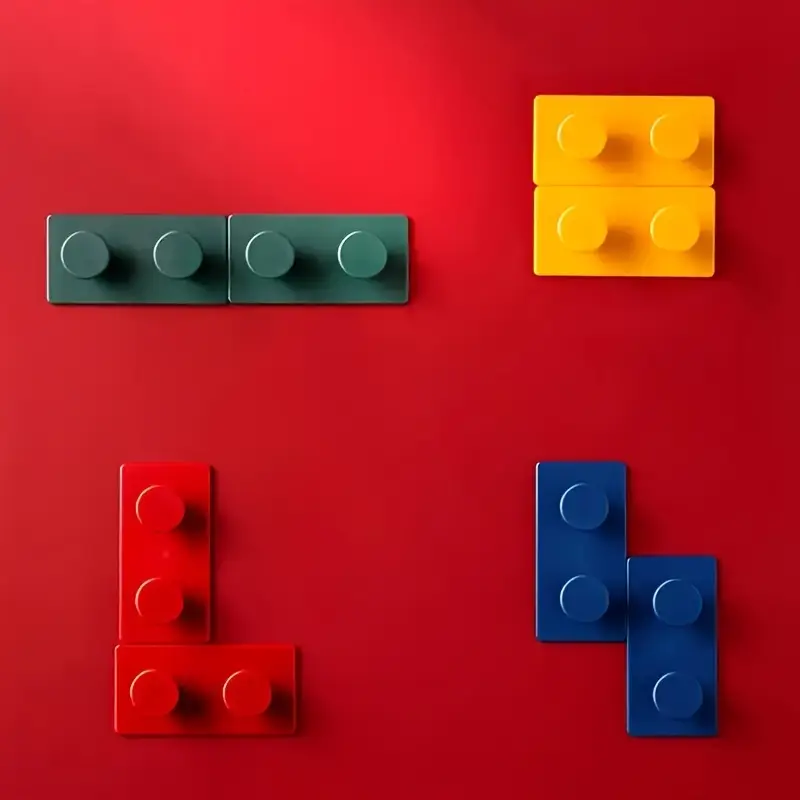 The 4-Pack Creative Building Block Wall Hooks, made from durable ABS resin, are artfully arranged on a red surface: green and yellow hooks on top, red and blue below, each in varying sizes and shapes.