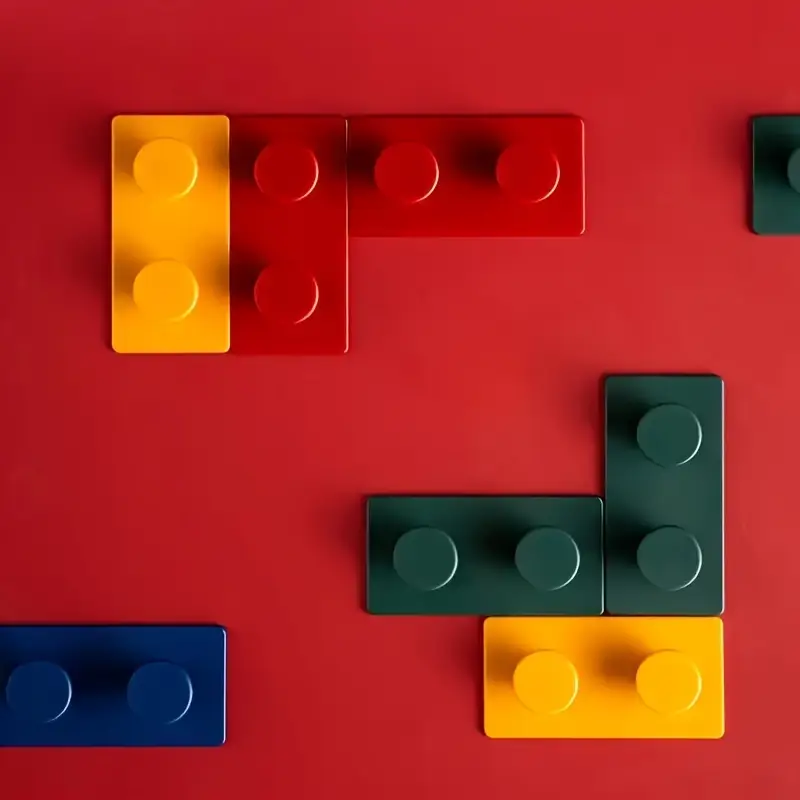 Yellow, red, green, and blue rectangular blocks made from durable ABS resin, featured against a red backdrop. Each block has two raised circles on top forming an abstract pattern that evokes the design of the 4-Pack Creative Building Block Wall Hooks.