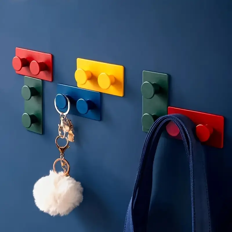 The 4-Pack Creative Building Block Wall Hooks, made from durable ABS resin, feature strong adhesive grips to hold items like a white fluffy keychain and blue bag strap. These vibrant storage hangers come in red, yellow, blue, and green, adding a playful touch to any space.