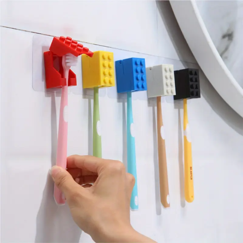 A hand selects a pink toothbrush from a row of five, each neatly held by a Kids Childrens Toothbrush Holder Wall-Mounted Brick Blocks Style. The durable plastic holders come in assorted colors: red, blue, white, yellow, and black.