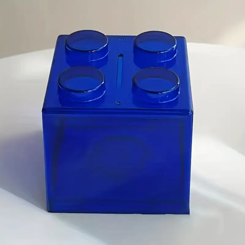 A blue, toy brick-like Building Block Piggy Bank with a top slot and four circular studs rests on a white surface. Soft shadows surround it, making it perfect for cultivating saving habits in a fun way.