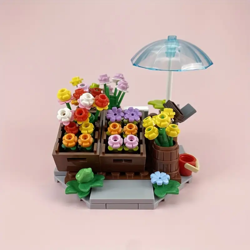 The Minifigure Flower Cart Plant Bouquet Building Blocks Set creates a lively scene with vibrant flowers in crates, a blue umbrella, and a watering can. A small frog sits on a stone platform, capturing the essence of an enchanting European Town Florist Stand against a soft pink background.