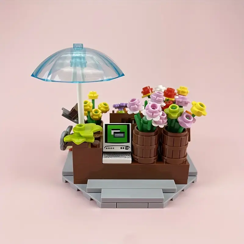 The Minifigure Flower Cart Plant Bouquet Building Blocks Set features a charming flower shop with colorful flowers, a small computer register, an umbrella, and includes a delightful minifigure flower cart for added play possibilities. Set on a gray base with a pink background.