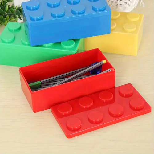 A red pencil case from a Pack of 4 2x4 Storage Blocks, shaped like interlocking blocks, has its removable lid open, showing pens and pencils inside. In the background, the yellow, blue, and green block-like storage blocks are closed.