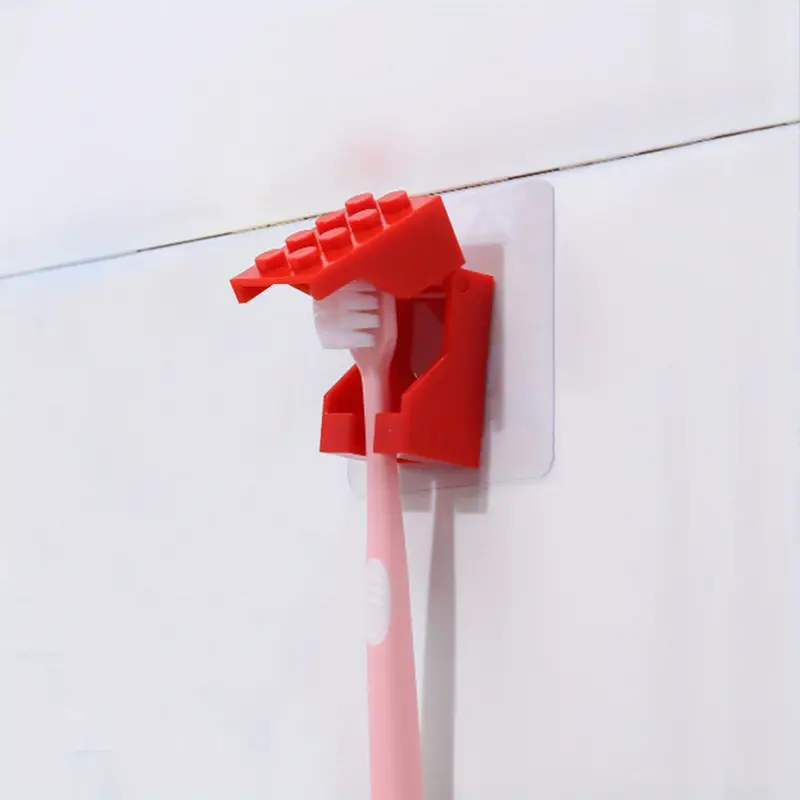 The Kids Childrens Toothbrush Holder, in a durable hand shape and brick block style, grips a pink toothbrush against the wall. This dustproof red holder uses a clear adhesive pad for seamless mounting on the white backdrop.