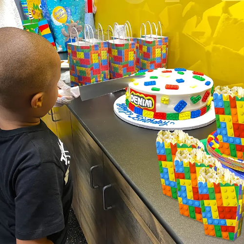 A child stands before a birthday cake with colorful block patterns, accompanied by whimsical party decor featuring 4 Themed Building Blocks Party Popcorn & Candy Boxes against a vibrant yellow wall.