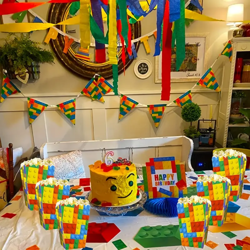 A vibrant birthday party features Lego-themed decorations with a yellow cake, 6 candle, colorful flags, streamers, and a cheerful Happy Birthday sign. The festive scene is completed by 4 Themed Building Blocks Party Popcorn & Candy Boxes for the perfect touch.