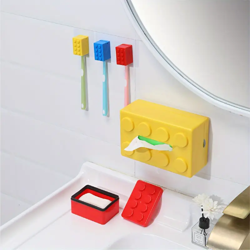 A bathroom showcases wall-mounted Kids Childrens Toothbrush Holders styled as red, blue, and yellow building block bricks made of durable plastic. Alongside, a yellow block-themed tissue box sits, with a small tray and vase on the sink complementing this playful ensemble.