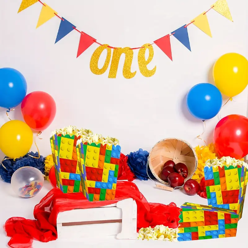 Festive party scene with 4 themed building blocks popcorn boxes, red and blue balloons, apples, and a small basket. A one banner hangs with gold letters and red, blue, yellow flags. A red cloth drapes over a white bench.