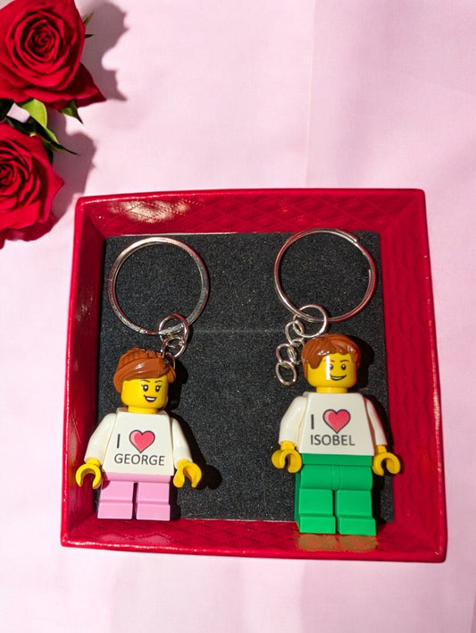 The Personalised LEGO® Valentines Keyring Minifigures for a couple set features two minifigure keychains in a red box with I ❤️ George and I ❤️ Isobel shirts, alongside red roses on a pink surface—a unique, personalized gift ideal for Valentines Day.