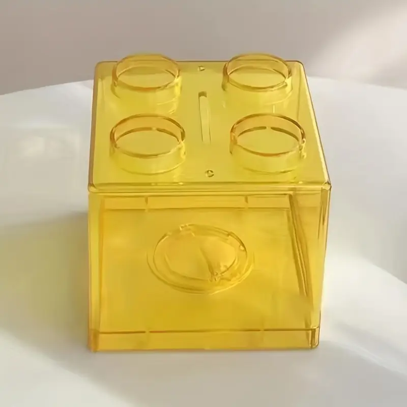 The transparent yellow box, resembling a large toy brick with four round protrusions on top, is actually the Building Block Piggy Bank. Ideal for cultivating saving habits, its designed to hold spare change while creating playful shadows on light surfaces.